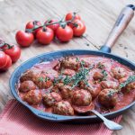 Albondigas (Fleischbällchen) in Tomatensauce mit Thymian in gußeiserner Pfanne mit Gabel, frischen Strauchtomaten und Knoblauch im Hintergrund. Gekocht nach Originalrezept von Papa Corazón Catering, Oberursel